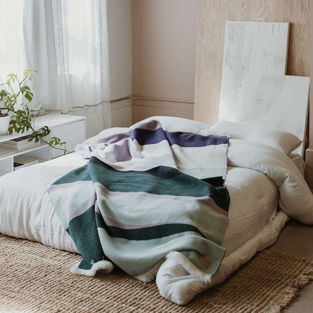 Green, teal, white and purple blanket lays on a bed with white linens